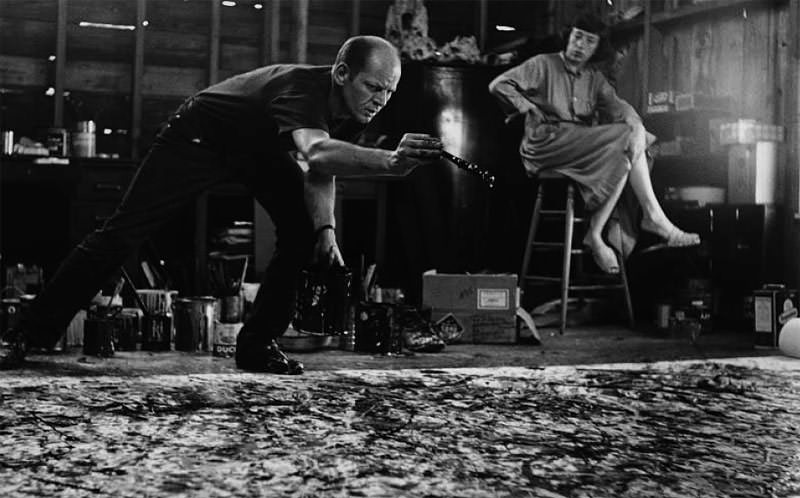 A black and white photo of Jackson Pollock using a paintbrush to splatter paint over a large canvas lying on the floor.